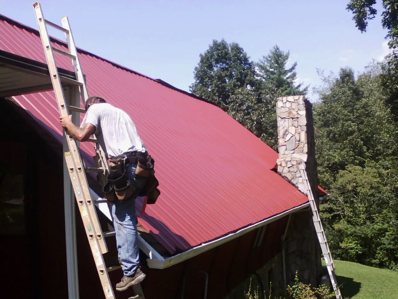 Roofer on a ladder beside a red metal roof and stone chimney — Soffit & Fascia Repair by Roofing Brothers in Eatonville